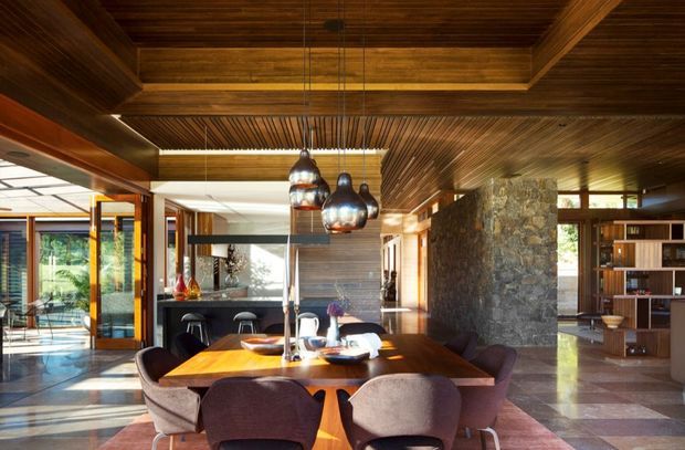 Open-plan dining and living area with a large timber dining table and upholstered chairs beneath sculptural pendant lights. The space features extensive dark timber ceiling panelling and beams, a rough natural-stone feature wall, polished stone or tiled flooring, expansive glazing, and built-in shelving. The room has a warm, refined mid-century-inspired character with strong indoor-outdoor connections.