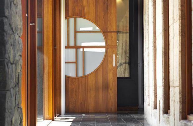 A close view of a refined residential entryway featuring a prominent warm timber front door with geometric frosted-glass inserts and curved detailing. The entry has dark tiled flooring, natural stone walls, timber framing, and a black-framed glazed door or window visible beyond. Strong sunlight highlights the crafted joinery and contrasting masonry.