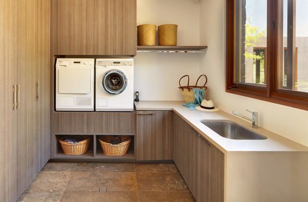 A compact, well-organised laundry room with warm wood-grain cabinetry, integrated white washer and dryer, open shelving, wicker baskets, a pale benchtop, stainless-steel sink, and a timber-framed window. The room has a practical L-shaped layout and earthy tiled flooring.
