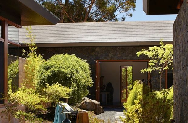 A landscaped courtyard entry framed by low, modern buildings. The facade combines dark rough stone walls, warm timber-framed glazing and doors, and a shallow light-grey tiled roof. Dense clipped shrubs, small trees, boulders, and a gravel path create a secluded, natural garden setting.