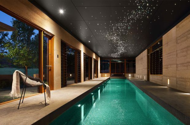 A dramatic indoor pool hall with a long turquoise swimming pool, dark star-like illuminated ceiling, warm timber-framed floor-to-ceiling glazing, and textured pale stone walls. The space opens toward landscaped outdoor views and includes a minimalist lounge chair, creating a luxurious resort-like atmosphere.
