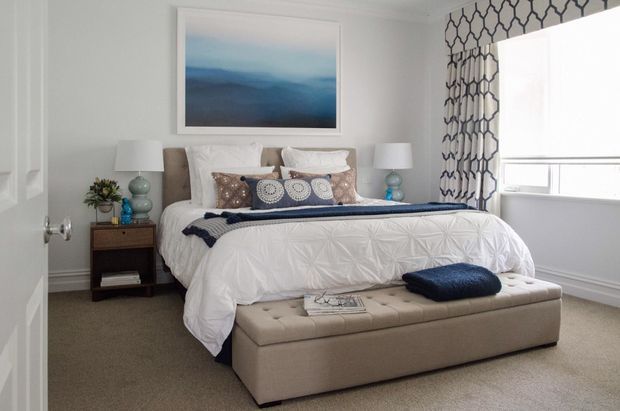 A bright, refined bedroom with a neatly made bed, upholstered bench at the foot, matching bedside tables and lamps, blue-and-white bedding, and a framed blue abstract artwork. White walls and trim, patterned window curtains, and beige carpet create a calm, polished interior.