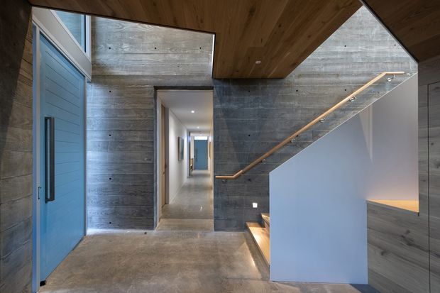 A contemporary interior entry hall with a dramatic stairway. The space features exposed horizontal concrete walls, a polished concrete floor, warm timber ceiling and soffit, a blue pivot entry door, recessed lighting, and a minimalist staircase with a clear glass balustrade and timber handrail. A corridor extends beyond the entry, while integrated stair and wall lighting adds warmth.