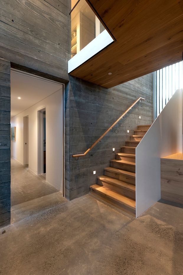 Interior view of a contemporary stairway and hallway. The stair rises beside a dark horizontally boarded feature wall, with warm timber treads, handrail, and soffit. A white balustrade, recessed stair lighting, polished concrete flooring, and a double-height void with upper-level glazing create a modern, warm industrial atmosphere.
