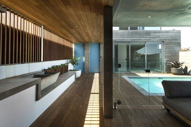 Covered modern outdoor living space beside a swimming pool, featuring warm timber decking and a timber-lined ceiling, a long built-in concrete planter bench, vertical timber screening, and expansive glass walls. The pool and adjacent concrete building create a clean, indoor-outdoor architectural composition with strong linear shadows.