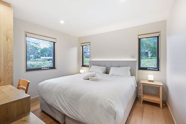 Bright, compact contemporary bedroom with a large upholstered bed dressed in white linen, light timber flooring, pale walls, black-framed windows, and simple timber bedside furniture. Built-in or fitted timber joinery is visible along the left wall, with greenery and neighbouring buildings visible through the windows.