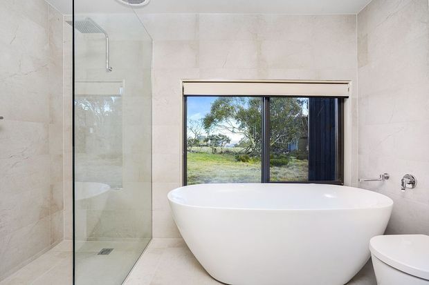 Bright contemporary bathroom featuring large pale stone-look wall and floor tiles, a frameless glass shower enclosure, a freestanding white oval bathtub, and a partially visible white toilet. A wide dark-framed window provides views of a rural landscape and natural light. The room has clean, minimal fittings and a calm neutral palette.