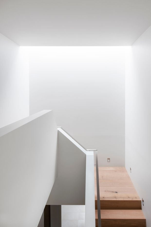 Minimalist interior view of a bright stairway with white plastered walls and balustrades, pale timber stair treads, and a high open stairwell. The composition emphasizes clean geometric planes, abundant diffuse light, and restrained detailing.