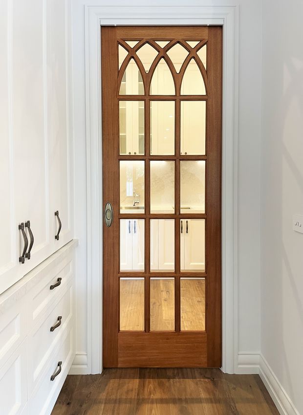 Interior view of a detailed timber-framed glazed door set within white decorative wall mouldings. The dark warm wood door features multiple glass panels and Gothic-inspired pointed arch tracery, with a white built-in cabinetry bank and dark timber flooring visible alongside it.