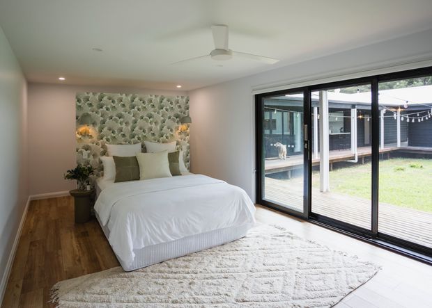 A bright contemporary bedroom with a low-profile upholstered bed dressed in white linens, muted green and cream cushions, and a textured botanical feature wall behind the bed. Warm timber flooring, a large neutral textured rug, recessed ceiling lights, and a ceiling fan create a relaxed feel. Full-height black-framed sliding glass doors open the room to a covered outdoor courtyard or deck.