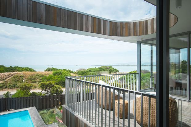 Contemporary coastal home exterior viewed from an elevated balcony or terrace, with curved timber-clad roof or soffit elements, extensive floor-to-ceiling glazing, a metal balustrade, and outdoor wicker seating. The view overlooks dense coastal vegetation and a broad body of water, while a small turquoise pool is visible below.