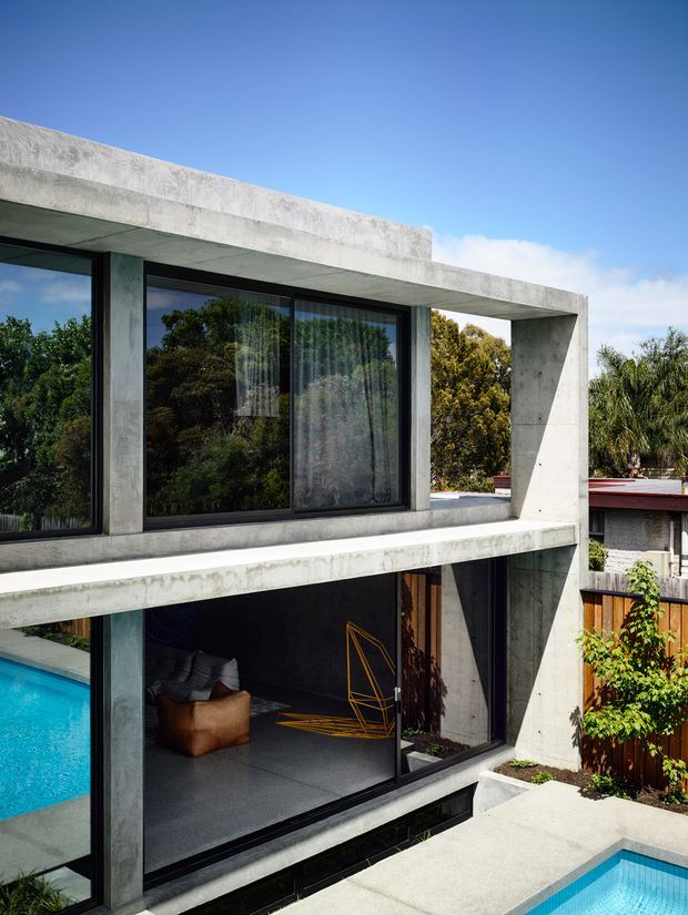 Exterior view of a contemporary two-storey concrete house with strong horizontal slabs, exposed concrete structure, dark-framed floor-to-ceiling glazing, and an adjacent swimming pool. The lower level opens toward a covered outdoor living area, while landscaped suburban surroundings are visible beyond the property.