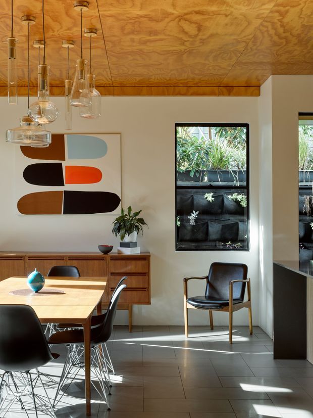 A warmly lit mid-century-inspired dining area with a timber dining table, black upholstered and molded chairs, a long timber sideboard, abstract artwork, and sculptural pendant lights. The room features a richly grained timber-panelled ceiling, pale walls, large dark-framed windows, and grey tiled flooring. A vertical planted garden is visible outside the windows.
