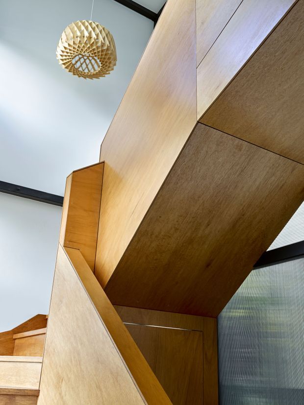 Upward-looking interior view of a sculptural timber stairway, with extensive warm wood panelling and angular geometric joinery. A woven wooden pendant light hangs from the high ceiling, while translucent ribbed glazing and dark structural beams appear along the right and upper edges.