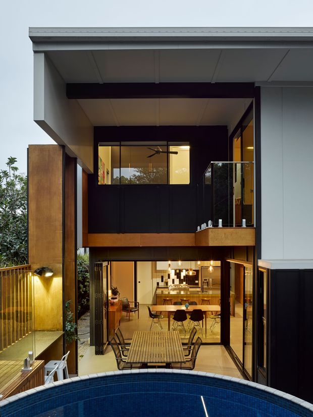 A contemporary multi-storey residential courtyard viewed from above a curved swimming pool. The home has a rectilinear form with projecting roof and upper-level balcony, dark charcoal wall cladding, pale metal roof and soffits, warm timber elements, extensive glazing, and an open-plan kitchen and dining area opening onto a timber deck. A narrow outdoor courtyard with planted edges and a small outdoor seating area is visible beside the house.