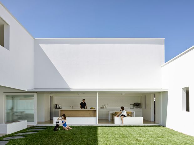 Bright contemporary courtyard exterior of a white, flat-roofed residential building. The view centres on an open-air entertaining area with a built-in outdoor kitchen or bar, white cabinetry, a timber-toned counter, and a covered dining zone. White rendered walls form a clean geometric composition around a grass courtyard, with strong sunlight and shadow across the facade and lawn.