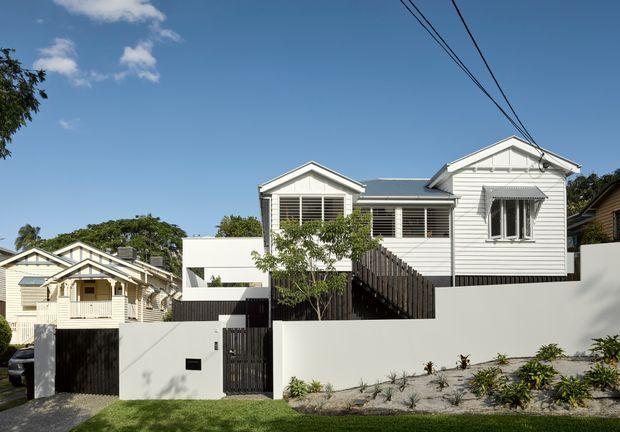 Exterior street view of a renovated, elevated timber-clad house with multiple gabled roof forms, white weatherboard walls, dark roofing, louvred windows, a timber staircase, and a white rendered boundary wall with landscaped planting. A neighbouring weatherboard house is partially visible to the left.