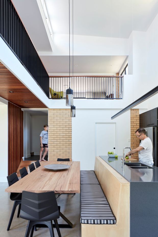 Bright, open-plan contemporary interior with a dining area and kitchen arranged around a double-height void. The space features a long light-timber dining table, black dining chairs, a kitchen island with pale timber cabinetry and a dark stone benchtop, integrated striped bench seating, exposed warm timber ceiling lining, pale brickwork, black vertical balustrades, and pendant lights. A mezzanine level and upper glazing emphasize the vertical volume.
