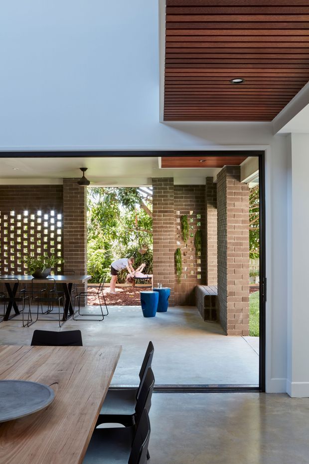 A contemporary indoor-outdoor dining and living area opening onto a landscaped courtyard. The space features exposed brown brick walls and columns, a polished concrete floor, expansive black-framed sliding glass doors, and a timber slatted ceiling with recessed lighting. A large timber dining table with black chairs occupies the foreground, while blue stools and built-in bench seating sit near the open courtyard. The architecture creates a strong connection between the interior and garden.