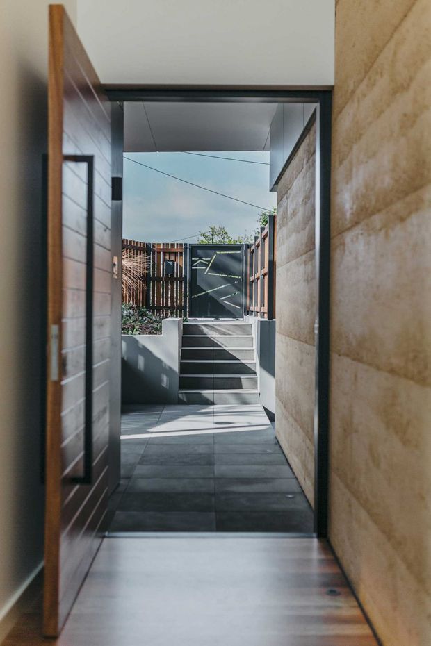 A narrow, modern entry passage viewed from inside, opening onto a small landscaped courtyard and a set of exterior steps. The space features dark grey floor tiles, a warm timber pivot door and wall lining on the left, a textured light stone wall on the right, dark metal door and window frames, and a sheltered flat-roof canopy. Strong sunlight creates geometric shadows across the paving and steps.
