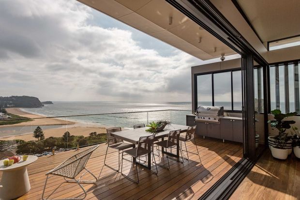 A bright coastal outdoor entertaining area viewed from inside through fully opened folding glass doors. The timber deck has a dining table, barbecue, lounge chair and potted plants, with expansive ocean and beach views beyond. The space features frameless or dark-framed glazed openings, a pale soffit, and a warm timber interior floor transitioning to the deck.