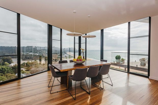 A bright contemporary dining area with a timber dining table and six upholstered chairs, set within floor-to-ceiling corner glazing. The room overlooks a coastal waterfront landscape with a terrace and railing visible outside. Warm timber flooring contrasts with slim dark window frames, a pale ceiling, and two suspended pendant lights.