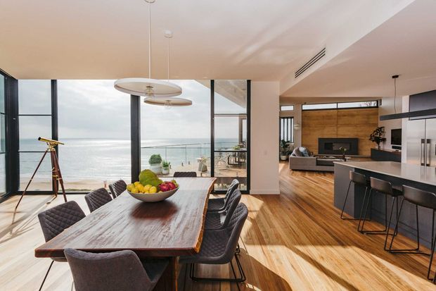 Open-plan contemporary interior combining a dining area, kitchen and living room, with full-height glazing opening to a balcony and expansive ocean views. The space features a long timber dining table, upholstered dining chairs, a streamlined island kitchen with dark cabinetry and stainless-steel appliances, timber flooring, recessed ceiling details, and a living area with a timber feature wall and fireplace.