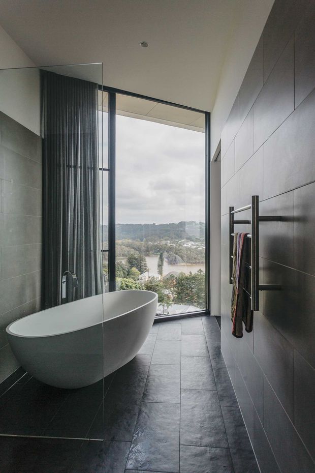 A sophisticated contemporary bathroom with a freestanding oval bathtub positioned beside a full-height glazed opening overlooking a leafy waterway landscape. Dark grey tiled flooring and large-format dark wall tiles create a moody, minimalist setting, with a frameless glass shower screen and wall-mounted heated towel rail visible.