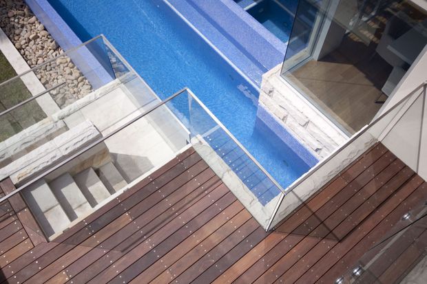 Elevated view of a modern outdoor terrace with dark timber decking, a narrow blue-tiled swimming pool, pale stone coping and steps, and clear glass balustrades. Large glazed doors open from the adjacent interior onto the deck, creating a seamless indoor-outdoor connection.