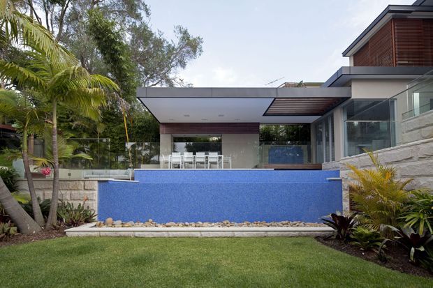 Contemporary multi-level residence viewed from the landscaped garden, featuring a vivid blue swimming pool, broad flat-roofed outdoor entertaining area, extensive glazing, horizontal roof planes, timber screening, and stepped stone retaining walls. Tropical planting and a manicured lawn frame the pool and house.