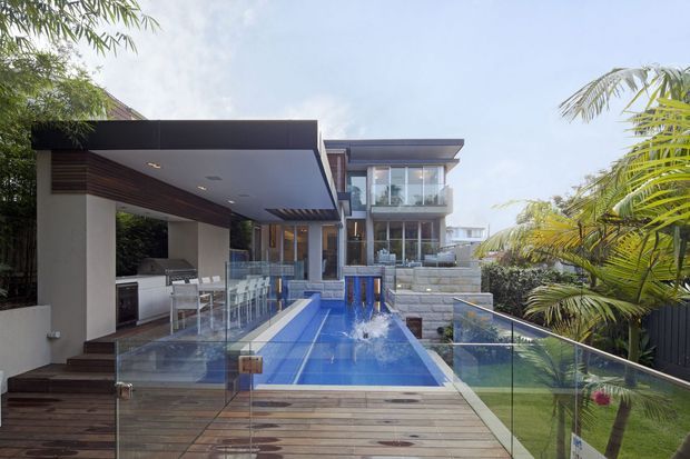 A contemporary multi-level residence viewed from an outdoor timber deck, featuring a narrow blue swimming pool with water jets, clear glass balustrades, a covered outdoor entertaining pavilion with built-in cabinetry, and landscaped tropical planting. The home has flat roof planes, extensive glazing, dark horizontal cladding accents, and pale masonry retaining walls.
