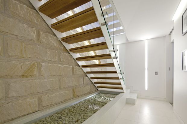 Bright contemporary interior stairway with open-riser timber treads, a clear glass balustrade, and a skylit void. A textured pale stone masonry wall runs alongside a decorative pebble-filled channel, while white tiled flooring and walls create a clean, minimalist finish.