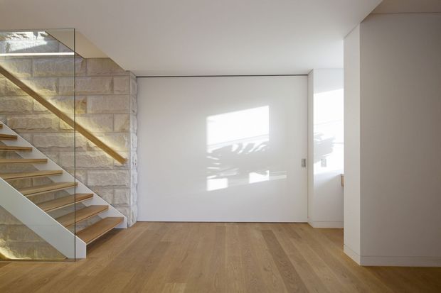 Bright minimalist interior hallway and stairway with warm timber flooring and open-riser timber stairs enclosed by a clear glass balustrade. A pale exposed masonry feature wall contrasts with smooth white walls, flush white doors, and clean-lined built-in detailing. Strong sunlight creates geometric shadows across the walls and floor.