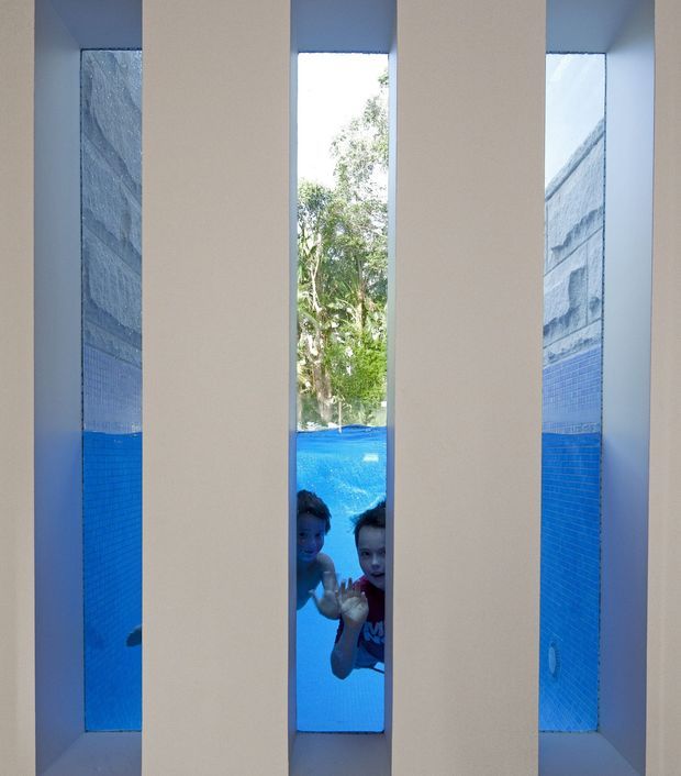 A narrow architectural view through vertical white wall openings onto a bright blue swimming pool. Two children are visible underwater near the central opening, with blue mosaic pool tiling and leafy greenery beyond. The image emphasizes the pool and the strong geometric rhythm of the wall structure.