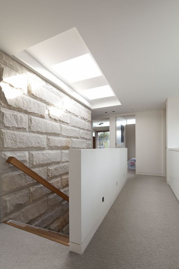 A bright contemporary residential landing or hallway at the top of a stairwell. The space features a white half-height balustrade, pale carpet, recessed ceiling lighting with three rectangular skylight-like panels, a substantial light rough-cut stone feature wall, and a simple timber stair handrail descending to the lower level. Glazed openings and a bedroom or adjacent room are visible beyond the landing.