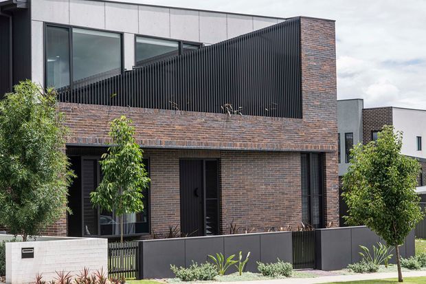 Contemporary multi-storey residential facade with dark brown brick walls, black vertical slatted balcony screening, dark-framed glazing, and a light grey upper wall plane. Low front fencing, landscaped planting, and neighbouring townhouses are visible.