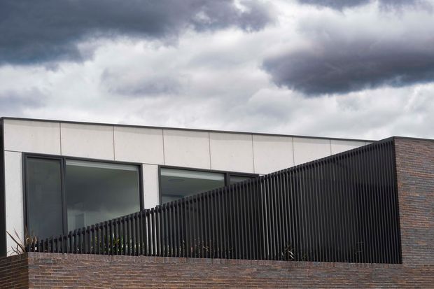 Exterior view of a contemporary, low-profile building with a long pale panelled facade, large dark-framed windows, a prominent black vertical-slat privacy screen, and a dark brick wall. Heavy overcast clouds create a subdued, dramatic atmosphere.