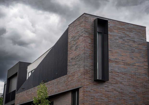 Exterior view of a contemporary multi-level building under a dramatic overcast sky. The facade combines extensive narrow-format reddish-brown brickwork with dark vertical metal screening, black-framed openings, and a tall slender projecting window. The composition emphasizes strong geometric lines and contrasting textures.