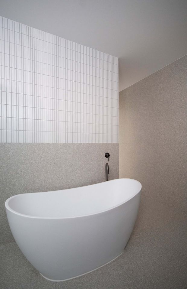 A minimalist bathroom featuring a freestanding white oval bathtub against walls finished in fine grey textured surfaces. The upper wall has narrow white vertical grid-pattern tiles, while a dark metal wall-mounted tap and slim bath filler provide contrast. The space has a restrained, contemporary material palette and clean geometric detailing.