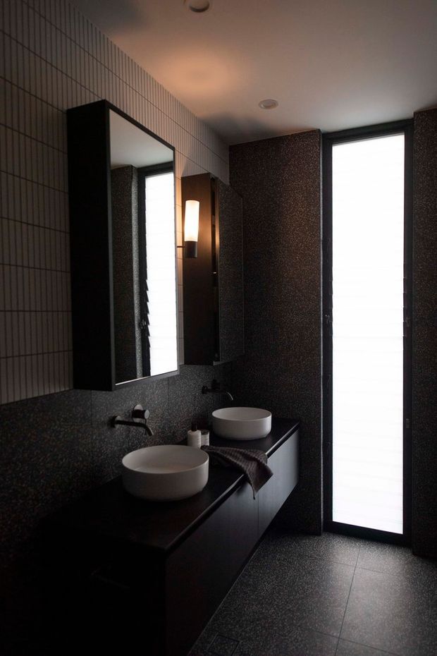 A dark, refined contemporary bathroom with a long black vanity, two white vessel basins, wall-mounted black taps, and a large framed mirror. The walls and floor are finished in dark speckled surfaces, while a vertical frosted-glass window or door brings in diffused light. A slim wall sconce provides warm illumination beside the mirror.