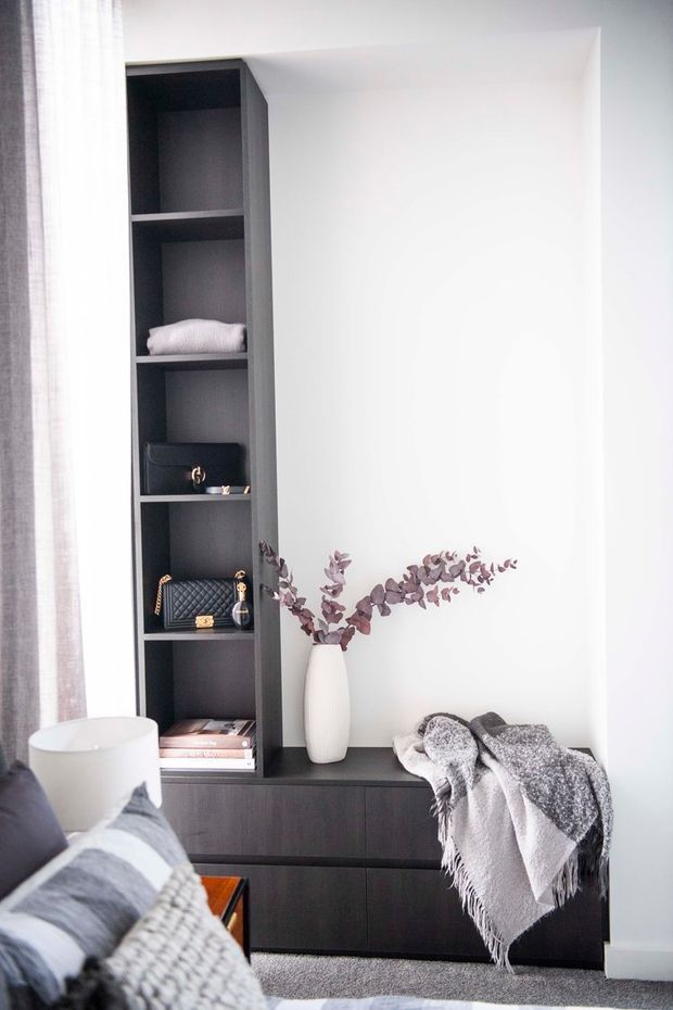 A bright, refined bedroom corner featuring a dark built-in shelving and drawer unit, styled with folded textiles, handbags, books, and a vase of dried foliage. Part of the bed with layered grey and white bedding is visible in the foreground beside a white table lamp. Soft curtains, white walls, dark joinery, and neutral textiles create a calm contemporary interior.