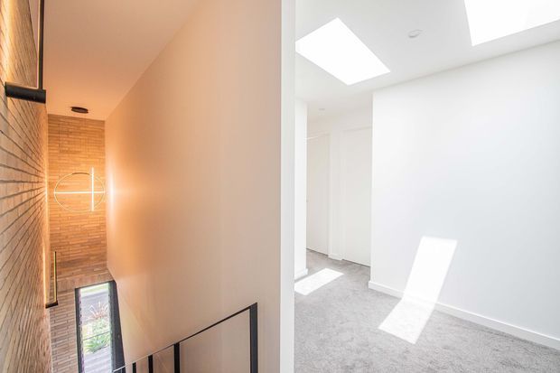 Split view of a contemporary residential interior: a warm-toned stairway with exposed brick walls, timber detailing, black metal balustrade and modern wall lighting on the left, alongside a bright white room with skylights, built-in cabinetry and grey carpet on the right.