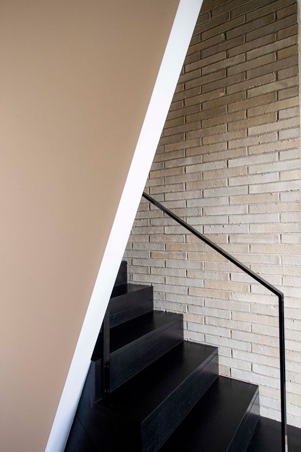 Interior architectural photograph of a compact staircase. Black timber or painted stair treads and risers rise alongside a pale exposed brick wall, with a slim black metal handrail. A sharply angled white stringer or wall edge creates a strong geometric diagonal in the foreground.