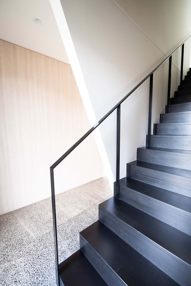 A minimalist interior stairway with dark timber or painted stair treads and risers, slim black metal balustrades, white walls and ceiling, and a pale vertical timber-panelled wall. The stair rises along the right side of the image, while a speckled terrazzo floor occupies the lower landing. A linear ceiling light creates a bright, clean architectural accent.