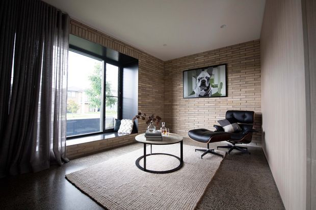 A refined contemporary living room with exposed light brick walls, dark-framed floor-to-ceiling glazing, dark curtains, and a neutral textured rug over a polished dark floor. The room is furnished with a black leather lounge chair and ottoman, a round black-framed coffee table, and a small side table. A framed artwork provides a monochrome focal point, while the large window brings in daylight and views of the outdoors.