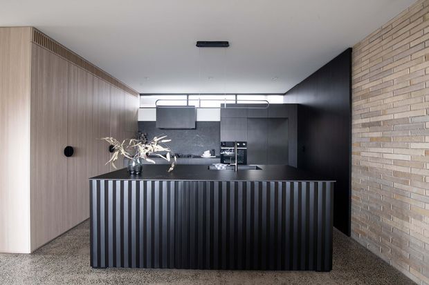 A refined contemporary kitchen viewed directly from the living area, featuring a large black island with dramatic vertical fluted detailing, dark built-in cabinetry, integrated appliances, and a pale timber-toned wall of cabinetry on the left. A horizontal clerestory window brings in natural light, while a textured light brick wall frames the right side. The floor appears to be a speckled terrazzo or polished aggregate finish, creating a monochromatic, high-contrast interior.