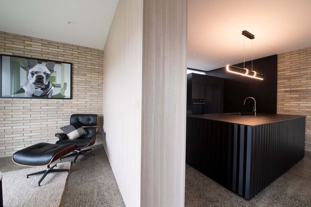 Contemporary open-plan interior divided by a vertical timber-toned partition, with a black kitchen and island on the right and a lounge area with a leather lounge chair and ottoman on the left. Pale exposed brick walls, dark cabinetry, a stone-look benchtop, carpeted flooring, and sculptural pendant lighting create a refined, high-contrast scheme.