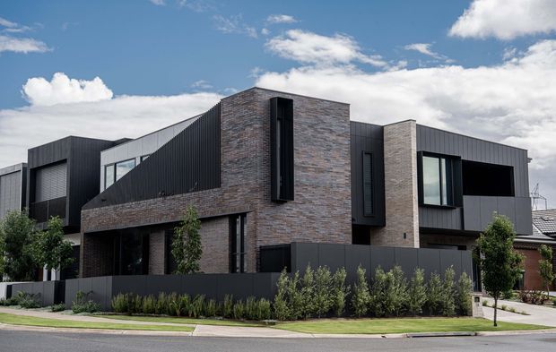 Street-facing exterior photograph of a contemporary multi-storey residence with a strongly geometric form. The facade combines dark vertical metal cladding with warm brown-grey brickwork, tall narrow windows, projecting box-like volumes, and a low dark boundary fence. Minimal landscaping sits between the building and the sidewalk under a bright blue sky.