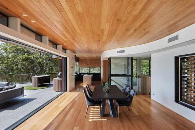 A bright, open-plan contemporary dining and living area with extensive glazing opening onto an outdoor terrace. The room features a long dark dining table with black chairs, warm timber flooring, a full-height timber-lined ceiling, white walls, built-in cabinetry, and a wall-mounted wine refrigerator. A compact kitchen and outdoor entertaining area are visible beyond the dining space.