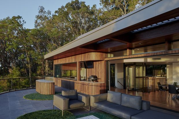 A contemporary single-level home set among dense eucalyptus woodland, photographed from an outdoor terrace. The building has a broad low-pitched roof with deep eaves, extensive glazing, warm vertical timber cladding, exposed timber soffits, and a covered outdoor entertaining area with lounge seating and a built-in barbecue. A paved terrace, curved timber-clad feature structure, and glimpses of the dining and kitchen areas are visible.